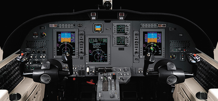 Aircraft cockpit featuring dual controls, multiple flight instruments, and navigation screens illuminated against a black background.
