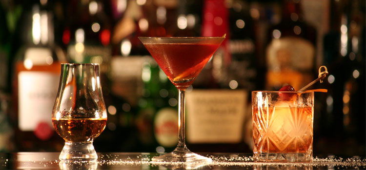 Bar display with a whiskey glass, a martini, and an old-fashioned cocktail surrounded by blurred bottles in the background.