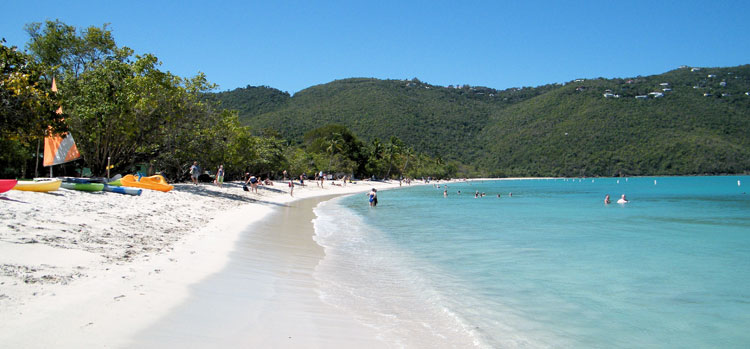 Tropical beach with clear blue water, white sand, kayaks, trees, and hills in the background under a sunny sky.