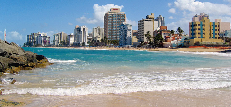 Urban beachfront with tall hotels and buildings next to clear blue water and golden sand under a bright sky.